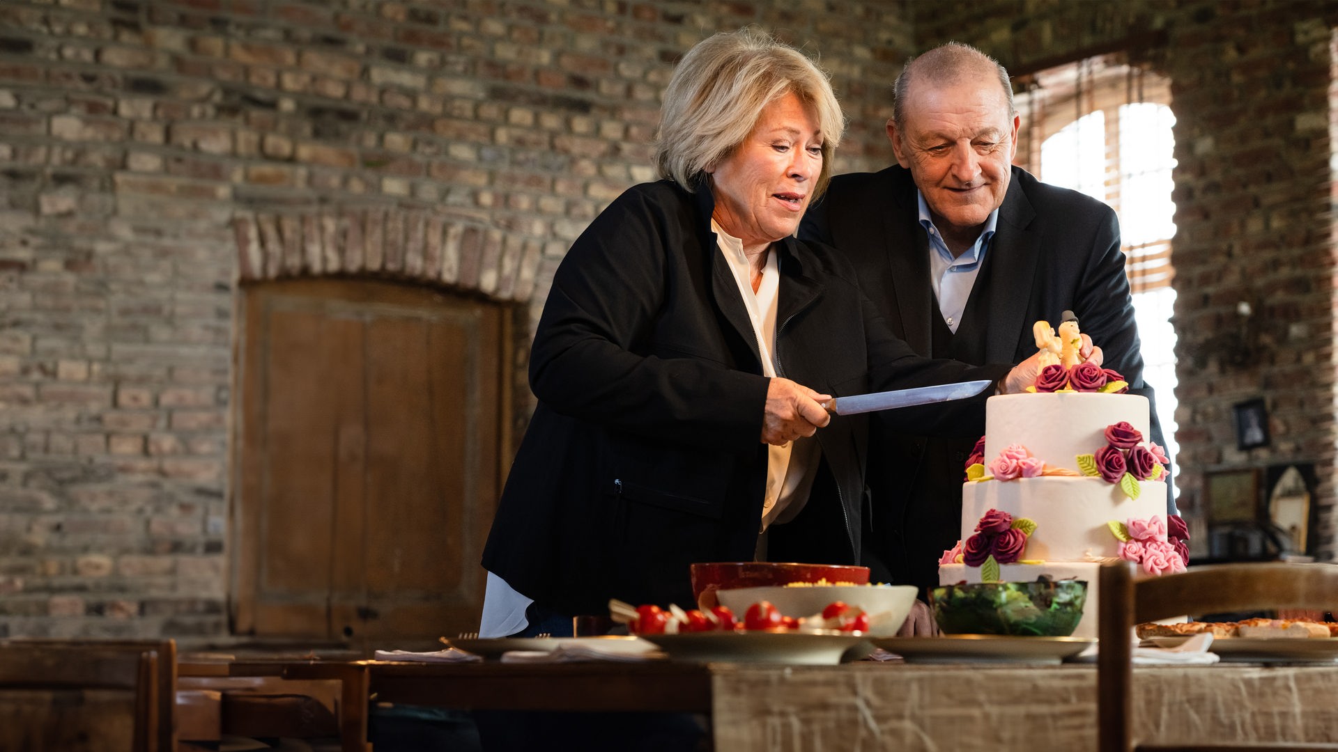 "Wilsberg - Einfach weg": Anna Springer (Rita Russek) und Wilsberg (Leonard Lansink) stehen zusammen in einer Wohnung vor einer mehrstöckigen, weißen Hochzeitstorte mit Rosen und Brautpaar. Anna hält ein Messer zum Schneiden der Torte hoch.