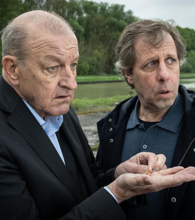 Wilsberg - Einfach weg": Wilsberg (Leonard Lansink) und Ekki (Oliver Korittke) stehen an einem Kanalufer. Wilsberg hält in seiner ausgestreckten Hand eine Goldkette.