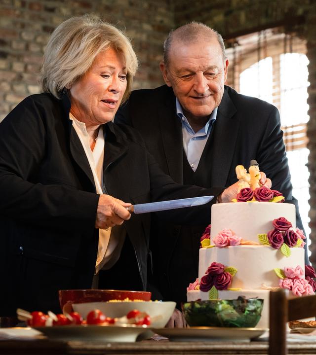 "Wilsberg - Einfach weg": Anna Springer (Rita Russek) und Wilsberg (Leonard Lansink) stehen zusammen in einer Wohnung vor einer mehrstöckigen, weißen Hochzeitstorte mit Rosen und Brautpaar. Anna hält ein Messer zum Schneiden der Torte hoch.