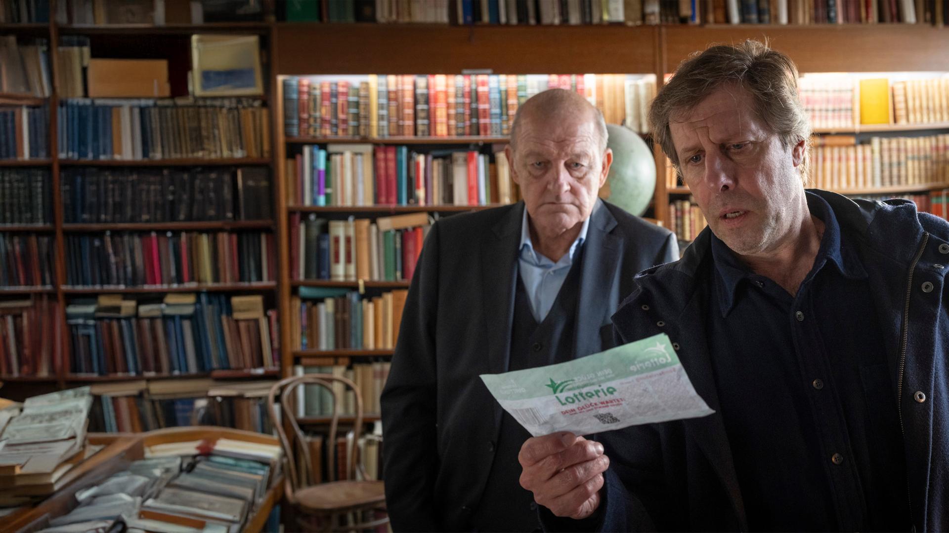 "Wilsberg - Todsicherer Tipp": Ekki Talkötter (Oliver Korittke) und Georg Wilsberg (Leonard Lansink) stehen nebeneinander im Antiquariat und blicken gemeinsam auf einen ausgefüllten Lotterieschein, den Ekki in der Hand hält.
