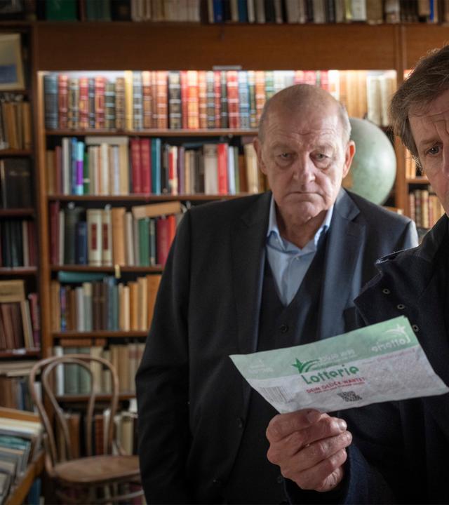 "Wilsberg - Todsicherer Tipp": Ekki Talkötter (Oliver Korittke) und Georg Wilsberg (Leonard Lansink) stehen nebeneinander im Antiquariat und blicken gemeinsam auf einen ausgefüllten Lotterieschein, den Ekki in der Hand hält.