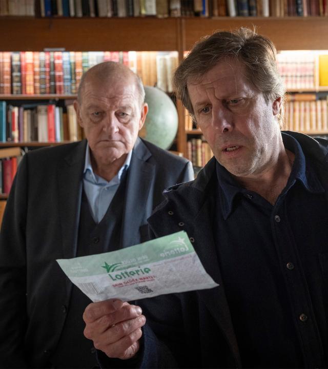 "Wilsberg - Todsicherer Tipp": Ekki Talkötter (Oliver Korittke) und Georg Wilsberg (Leonard Lansink) stehen nebeneinander im Antiquariat und blicken gemeinsam auf einen ausgefüllten Lotterieschein, den Ekki in der Hand hält.