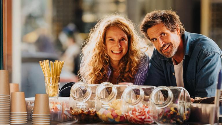 "Eine Wingwoman verliebt sich nicht": Caro Sander (Chiara Schoras) und Marc Ahmadi (Tom Beck) stehen nebeneinander an einem Kiosk. Vor ihnen stehen Süßigkeiten in Glasbehältern und Pappbecher. Beide schauen lächelnd nach vorne.