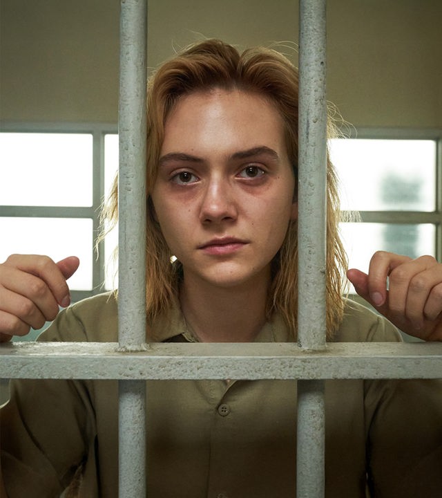 Reality (Emilia Jones) steht mit erschöpftem Blick, Augenringen und einem nahezu vollständig verheilten, blauen Auge hinter den Gitterstäben einer Gefängniszelle, an die sie sich mit ihren Handgelenken lehnt.