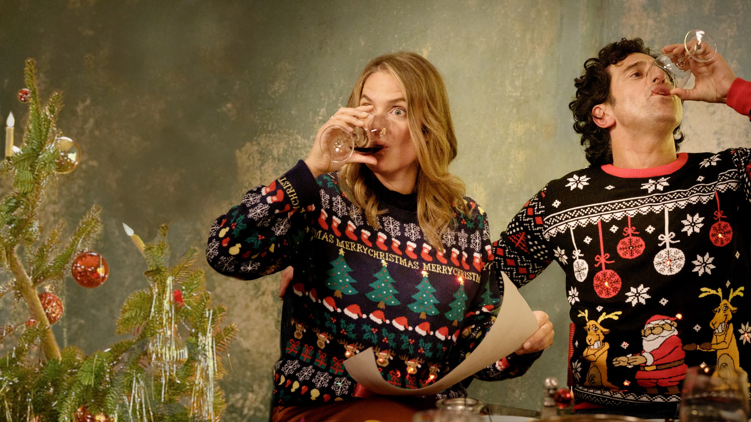 "Zwei Weihnachtsmänner sind einer zu viel": Karo und Lasse sitzen an einem Tisch und tragen bunte Pullover mit weihnachtlichen Motiven und LED-Lichtlein. Karo hält ein Blatt in der Hand. Beide nehmen einen kräftigen Schluck aus ihren Rotweingläsern.
