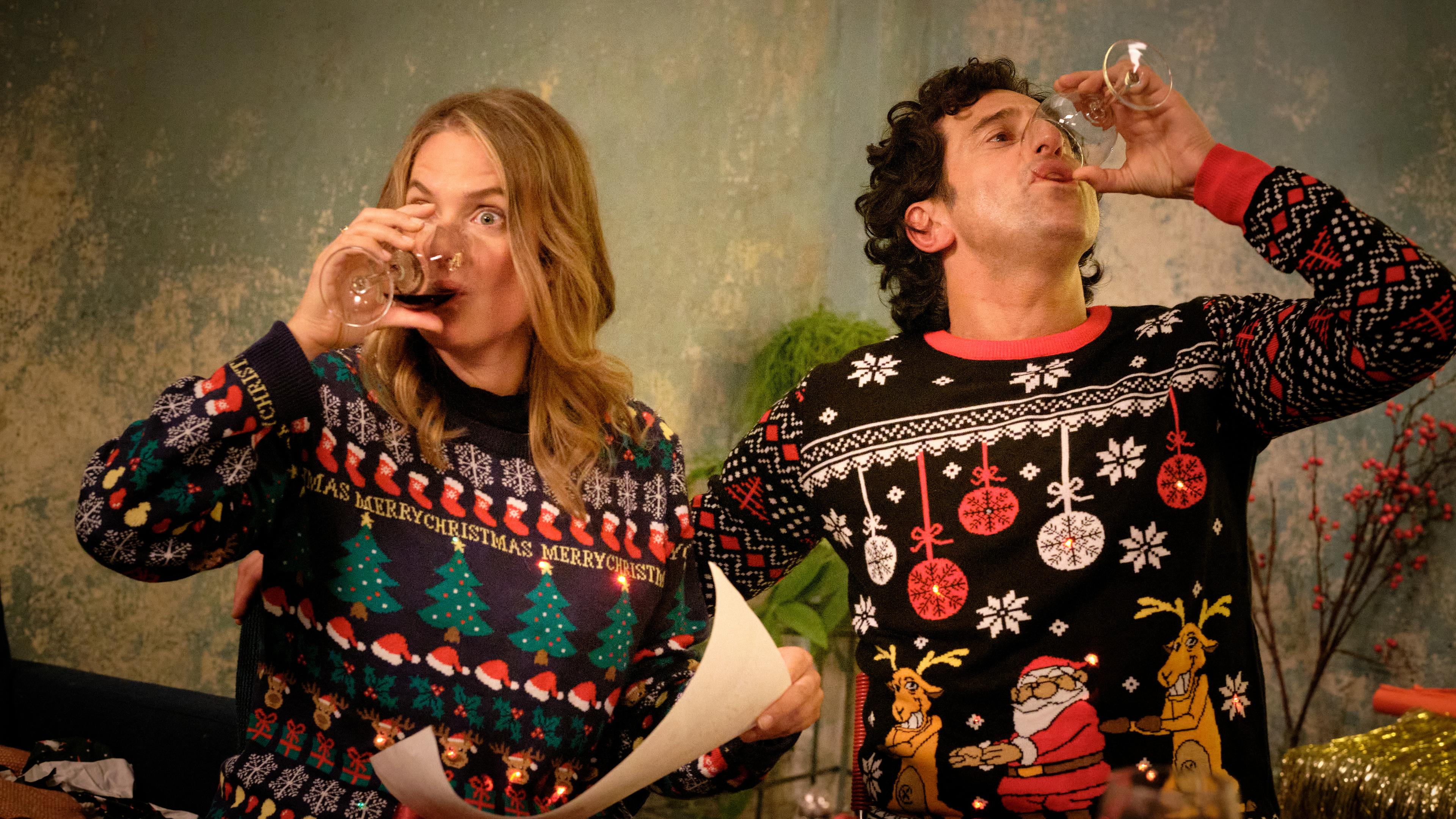 "Zwei Weihnachtsmänner sind einer zu viel": Karo und Lasse sitzen an einem Tisch und tragen bunte Pullover mit weihnachtlichen Motiven und LED-Lichtlein. Karo hält ein Blatt in der Hand. Beide nehmen einen kräftigen Schluck aus ihren Rotweingläsern.