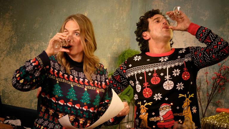 "Zwei Weihnachtsmänner sind einer zu viel": Karo und Lasse sitzen an einem Tisch und tragen bunte Pullover mit weihnachtlichen Motiven und LED-Lichtlein. Karo hält ein Blatt in der Hand. Beide nehmen einen kräftigen Schluck aus ihren Rotweingläsern.