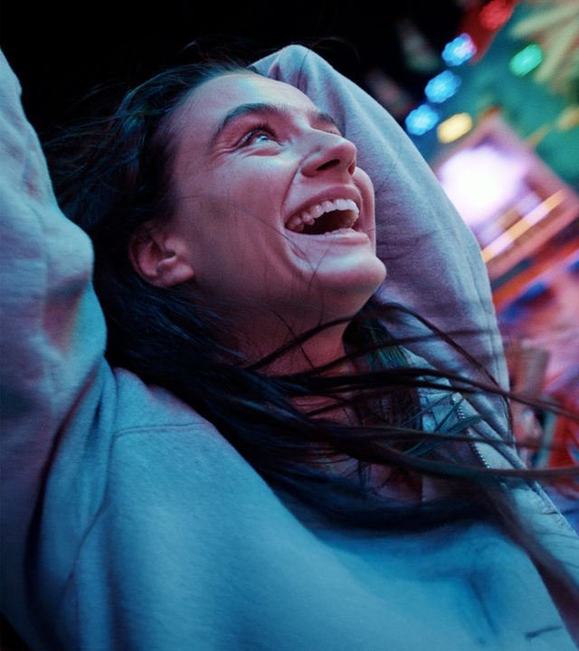 Marie (Laura Balzer) lacht glücklich und hat die Arme nach oben gehoben. Ihre Haare fliegen wild umher. Im Hintergrund sind die bunten Lichter des Jahrmarkts zu sehen.