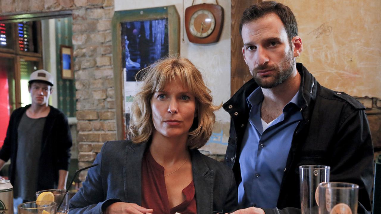 "Die Spezialisten - Im Namen der Opfer: Heroes": Dr. Katrin Stoll (Valerie Niehaus) und Henrik Mertens (Matthias Weidenhöfer) stehen bei Tag an der Bar eines Clubs. Vor ihnen liegt eine Akte, auf der das Bild einer Frau ohne Haare zu sehen ist. Stoll und Mertens blicken ihr Gegenüber, das nicht sichtbar ist, ernst an. David Hehling (Vincent Krüger) steht im Hintergrund und hört dem Gespräch zu.