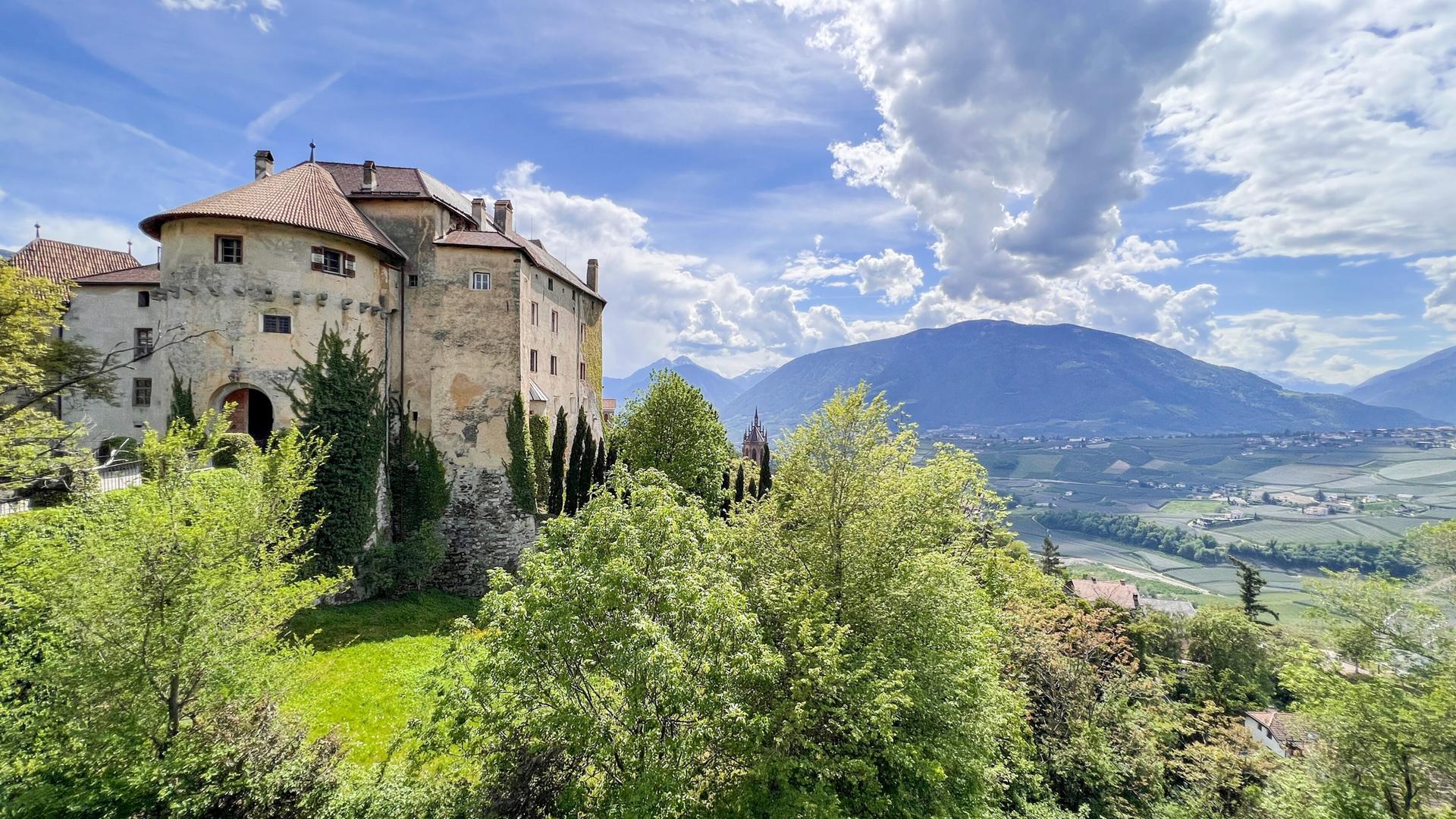Schloss Schenna thront auf einem Hügel über Meran, umgeben von Bäumen und mit Blick auf die Berglandschaft Südtirols.