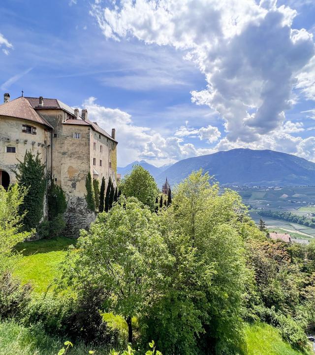 Schloss Schenna thront auf einem Hügel über Meran, umgeben von Bäumen und mit Blick auf die Berglandschaft Südtirols.