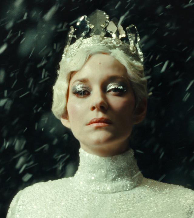 Porträtfoto einer weißblonden Frau mit silbrig glitzerndem Diadem in einem engen silbgrig glitzernden Rollkragen-Oberteil mit dickem silberfarbenem Lidschatten vor einem dunklen Hintergrund.