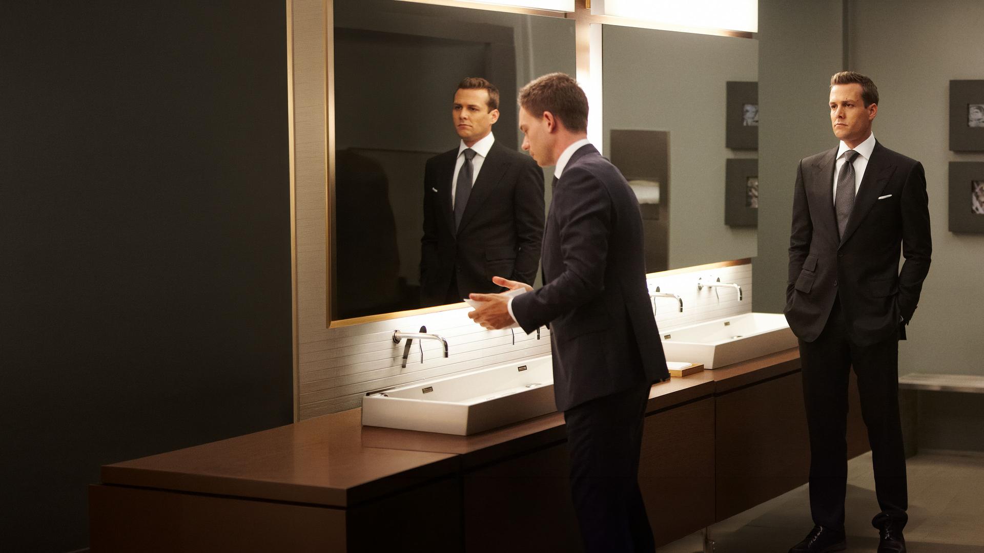 "Suits - Herzenssache": In einem Toilettenraum: Mike (Patrick J. Adams) steht an einem Waschtisch vor einem großen Spiegel, während Harvey (Gabriel Macht) mit ernster Miene im Raum steht und ihn beobachtet.