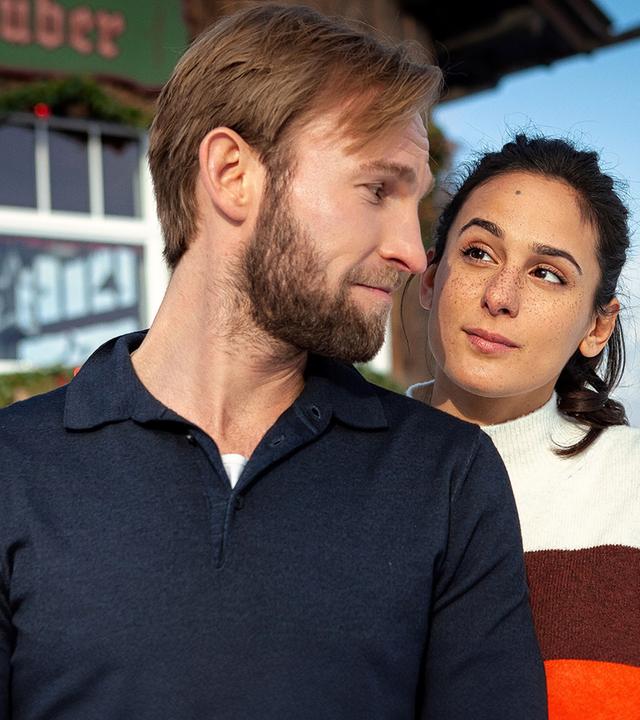 Jakob (Jochen Matschke) mit einem blauen Pullover und Alice (Aybi Era) mit einem gestreiften Pullover stehen dicht nebeneinander vor einem Holzhaus. Er blickt sie über die Schulter herausfordernd an, sie hat skeptisch eine Augenbraue hochgezogen. Es liegt Schnee und die Sonne scheint.