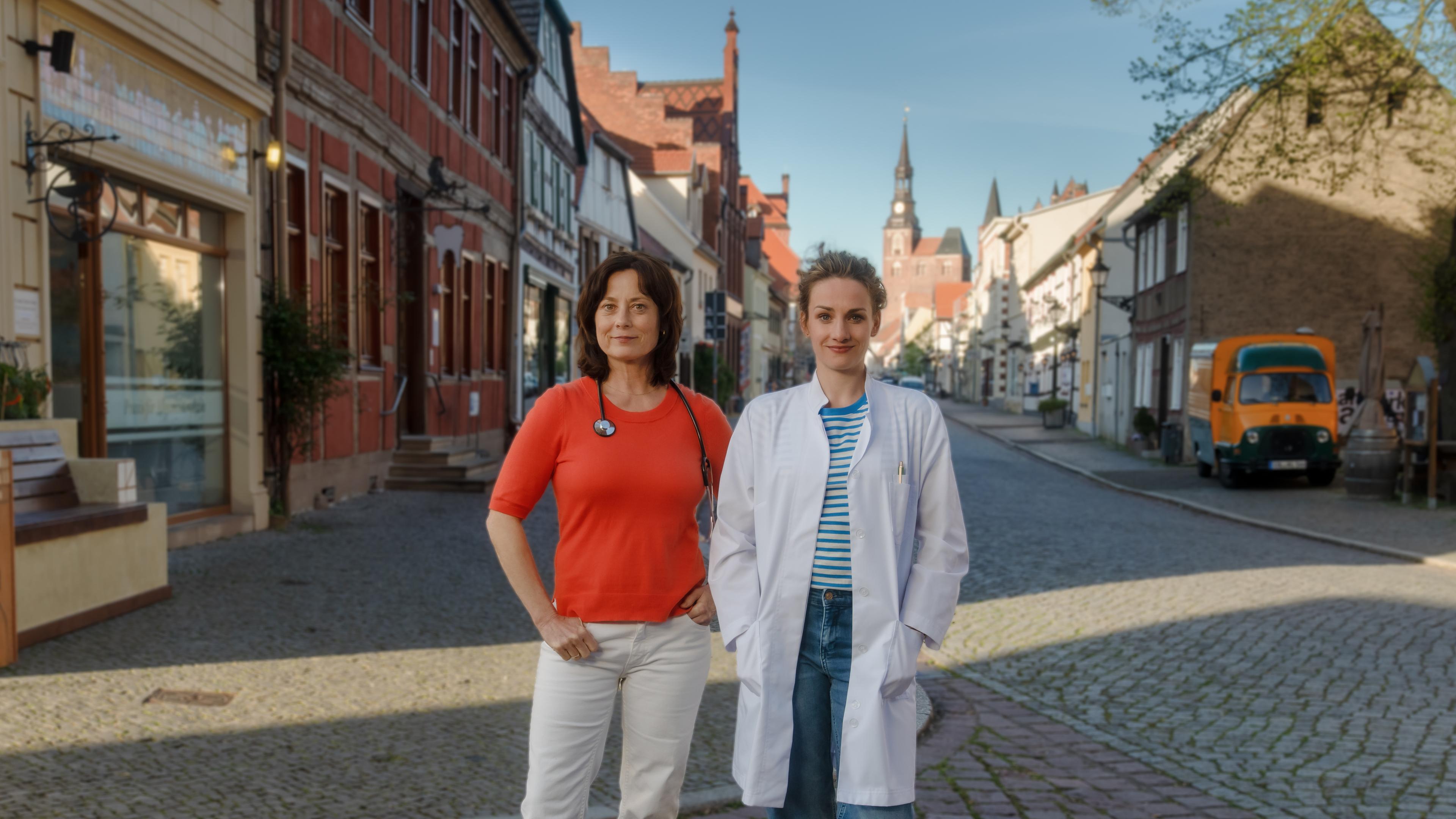 Neben der Arztpraxis stehen Dr. Katrin Herz (Inka Friedrich) und Dr. Holly Sass (Karoline Teska) auf der Straße. Katrin hat ein Stethoskop um den Hals gelegt, Holly trägt einen weißen Arztkittel. Beide blicken lächelnd in die Kamera.