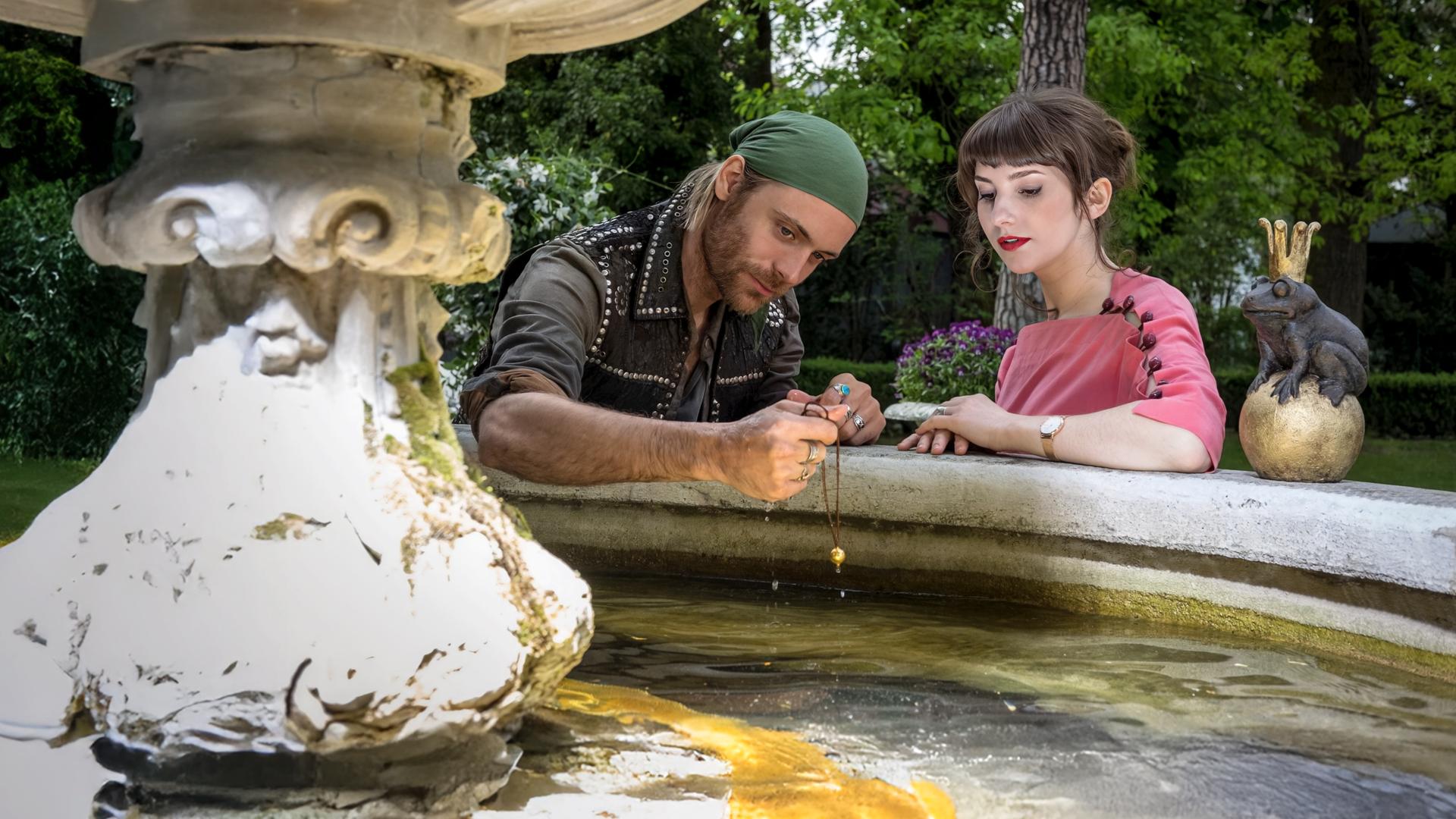 "Herzkino.Märchen: Der Froschkönig": Jimi (Bernd-Christian Althoff) und Isabelle (Anuschka Tochtermann) sitzen am Rande eines Brunnens, auf dem ein Frosch auf einer goldenen Kugel sitzt. Er hält eine Kette mit einer goldenen Kugel in der Hand.
