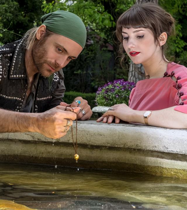 "Herzkino.Märchen: Der Froschkönig": Jimi (Bernd-Christian Althoff) und Isabelle (Anuschka Tochtermann) sitzen am Rande eines Brunnens, auf dem ein Frosch auf einer goldenen Kugel sitzt. Er hält eine Kette mit einer goldenen Kugel in der Hand.