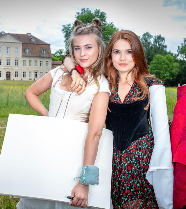 "Herzkino.Märchen: Schneeweißchen und Rosenrot": Die Schwestern Rosalie (Zoe Moore) und Bianca (Jeanne Goursaud) halten Zeichenblock sowie Stoffballen in den Armen und stehen auf einer grünen Wiese. Im Hintergrund ist das Schloss.