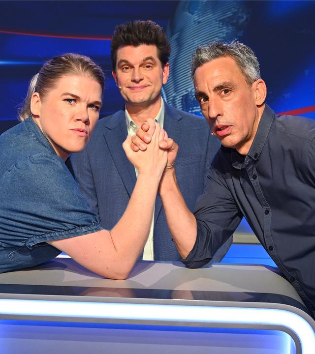 Gisa Flake (links) und Alexander Schubert (rechts) liefern sich im Studio ein Duell im Armdrücken. Zwischen ihnen befindet sich Quizmaster Lutz van der Horst.