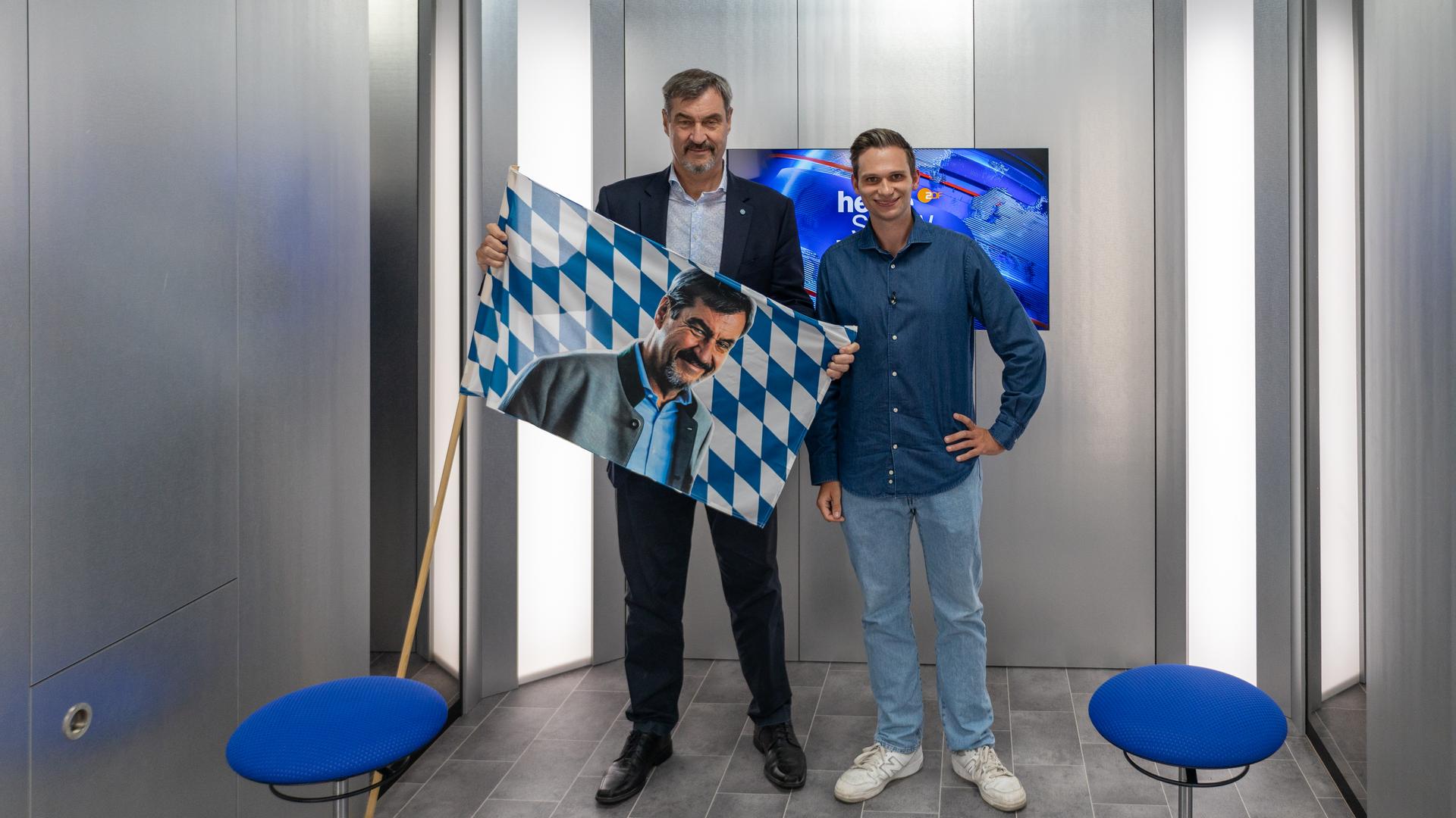 Markus Söder und Fabian Köster stehen in einem kleinen Raum. Söder hält die bayrische Flagge in den Händen, statt des Wappens ist jedoch ein Foto von ihm auf die Flagge retuschiert.