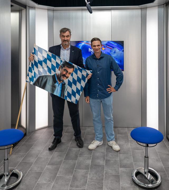 Markus Söder und Fabian Köster stehen in einem kleinen Raum. Söder hält die bayrische Flagge in den Händen, statt des Wappens ist jedoch ein Foto von ihm auf die Flagge retuschiert.