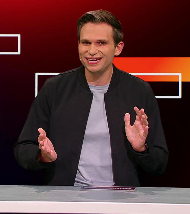 Fabian Köster sitzt in einem Studio an einem Schreibtisch. Links von ihm ist eine satirische Fotomontage von Friedrich Merz als Schiedsrichter zu sehen, der auf einen verletzten Fußballspieler zeigt, der auf dem Rasen liegt. Am unteren Bildrand steht "Faul!".