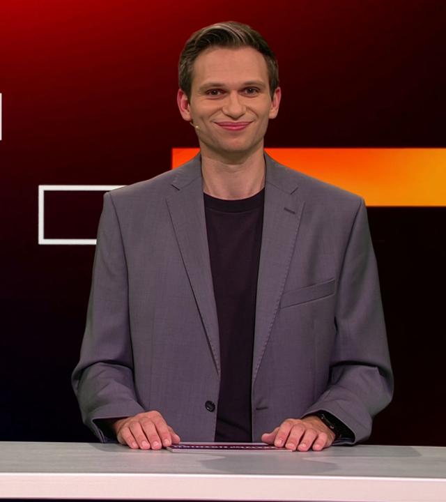 Fabian Köster sitzt in einem Studio an einem Schreibtisch. Links von ihm ist eine satirische Fotomontage von Alice Weidel und Tino Chrupalla zu sehen, die auf Wladimir Putins Schoß sitzen, während Putin aus einem Buch vorliest. Am unteren Bildrand steht "PUTIN ANTE PORTAS".