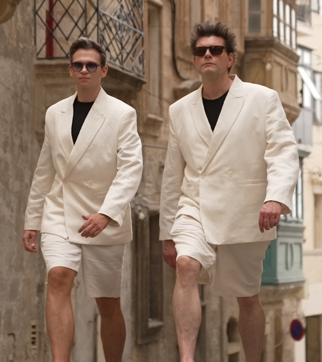 Lutz van der Horst und Fabian Köster tragen beide einen weißen Anzug und schwarze Sonnenbrillen und laufen durch eine Gasse. 