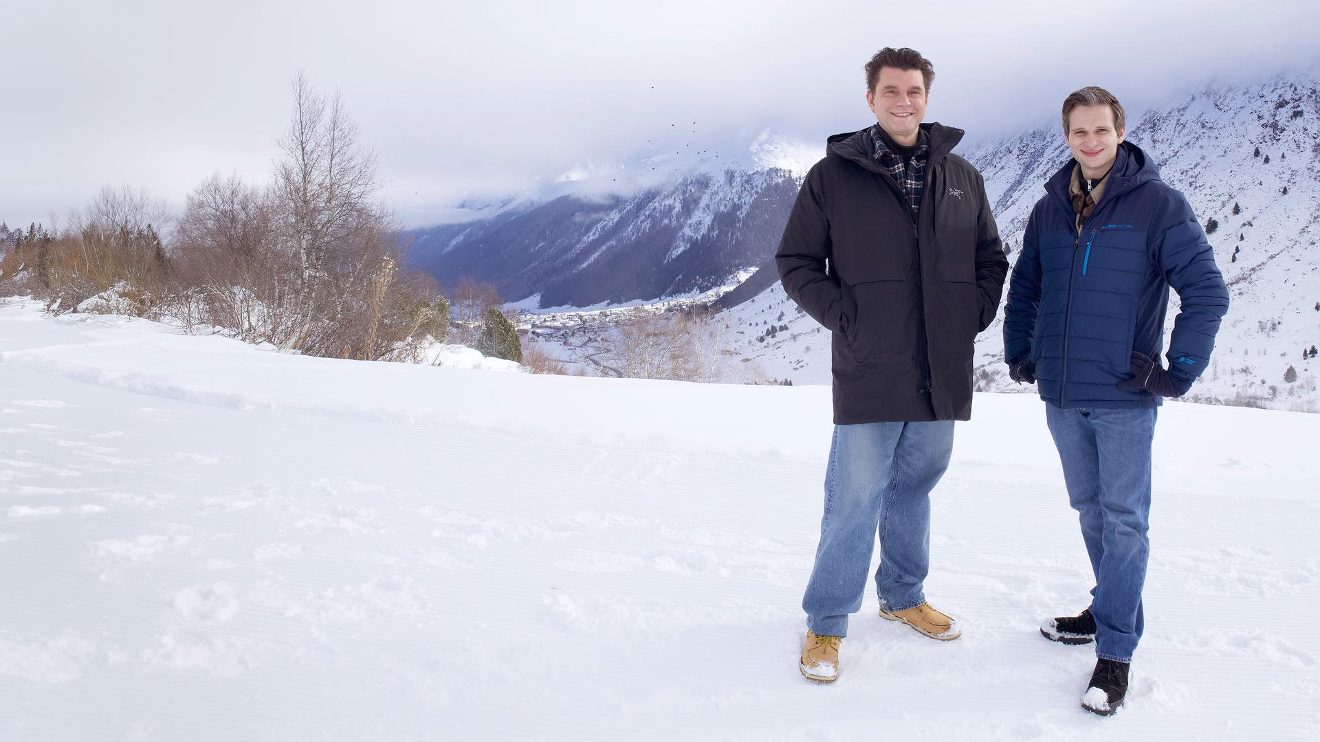 Lutz van der Horst und Fabian Köster stehen im Schnee des Skigebietes Galtür, im Hintergrund sind Teile der Silvretta-Gebirgsgruppe zu sehen.