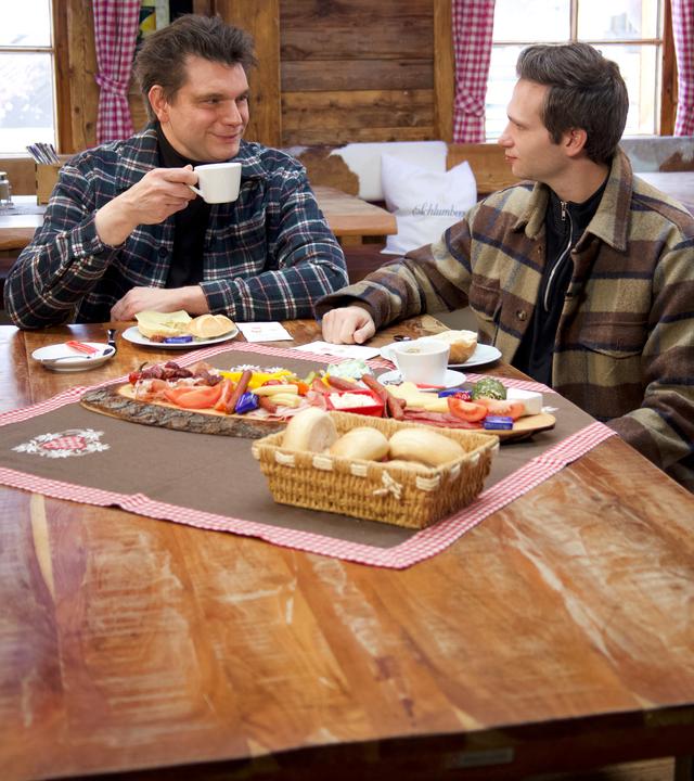 Lutz van der Horst (l.) und Fabian Köster (r.) sitzen an einem Tisch, der mit einer Brotzeit gedeckt ist.