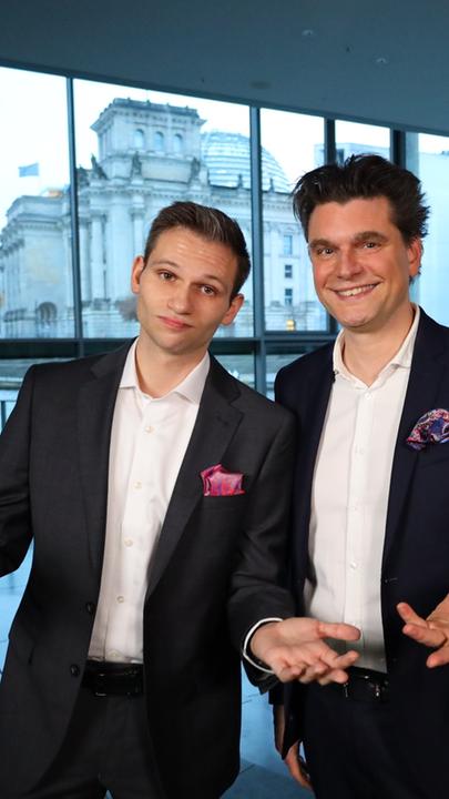 Fabian Köster und Lutz van der Horst stehen in einem verglasten Foyer und schauen freudig in die Kamera. Im Hintergrund ist das Reichstagsgebäude des Deutschen Bundestags in Berlin zu sehen.