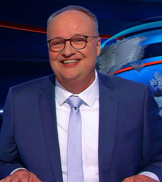 Oliver Welke sitzt rechts im Bild. Links im Bild ist eine satirische Fotomontage von Friedrich Merz in einem leeren Konferenzraum zu sehen. Vor ihm auf dem Tisch liegt ein kleiner Kuchen mit einer brennenden Kerze. Unter dem Bild steht "MERZ FANCLUB-TREFFEN".