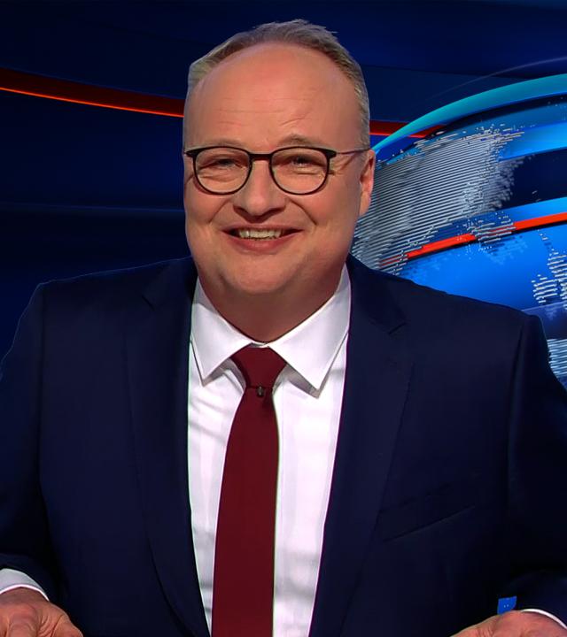 Oliver Welke sitzt am Schreibtisch. Auf der Bildkachel links neben ihm ist Friedrich Merz, vor dem ein Flakon der Marke "Boss" steht. Die Kachel trägt die Unterschrift "Der Duft, der Russen provoziert".