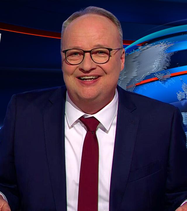 Oliver Welke sitzt am Schreibtisch. Links von ihm ist eine Fotomontage zu sehen, auf der Friedrich Merz und Angela Merkel abgebildet sind. Merkel hält sich ein kleines Fernglas vor die Augen. Unter dem Bild steht: "Ich kann nicht, wenn Mutti zuschaut".