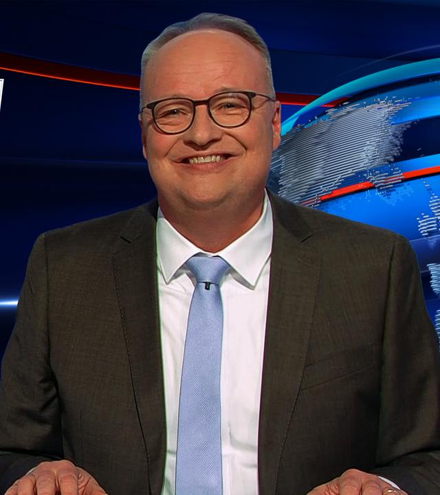 Oliver Welke sitzt rechts im Bild. Links ist eine satirische Fotomontage im Stil der Serie "Emily in Paris" zu sehen: Emily mit einem Fuß auf der Motorhaube eines brennenden PKW hält einen Benzinkanister. Darunter steht: "EMILY IN PARIS zündet Autos an".