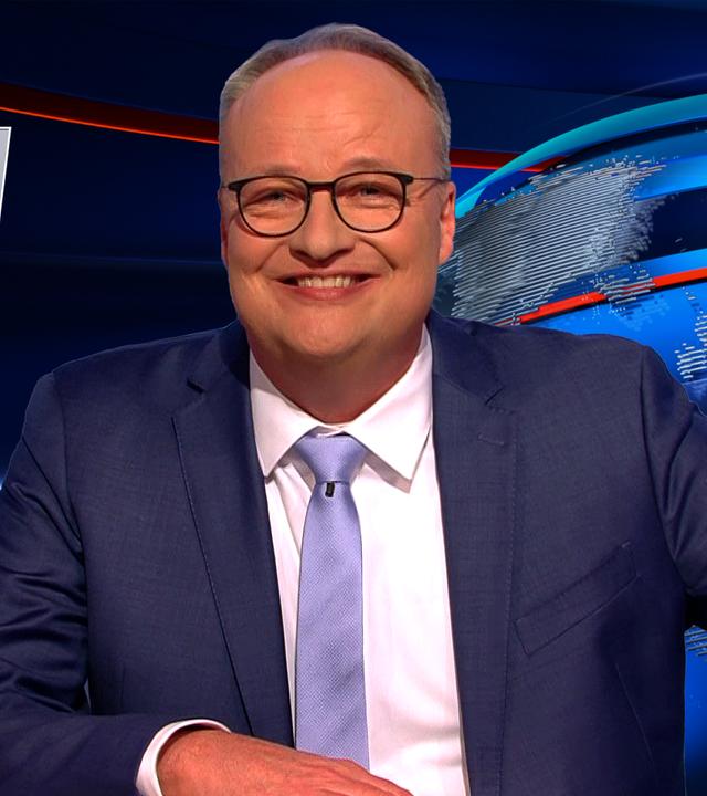 Oliver Welke sitzt am Schreibtisch. Links im Bild befindet sich eine Fotomontage von Friedrich Merz, der eine große Schultüte hält. Am unteren Bildrand steht der Text: "Meine erste Kanzlerschaft".