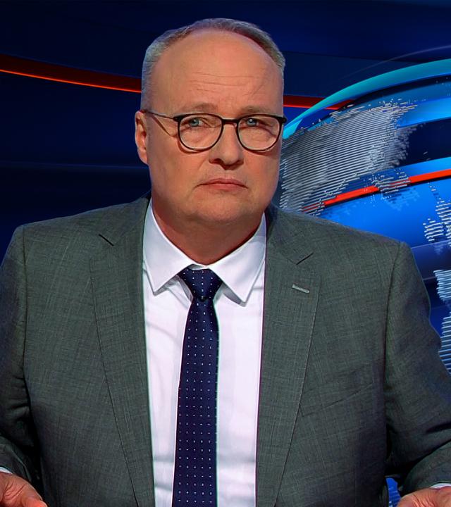 Oliver Welke sitzt rechts im Bild. Links ist eine satirische Fotomontage im Stil von "Die drei Fragezeichen" zu sehen. Über einem Foto von Ulrich Siegmund steht "Die vier Fragezeichen und die eingestürzte Brandmauer".
