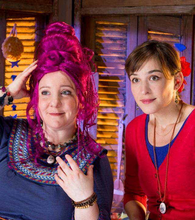 Elsebö steht mit einer verrückten Perücke vor dem Spiegel, neben ihr steht staunend Siebenstein.