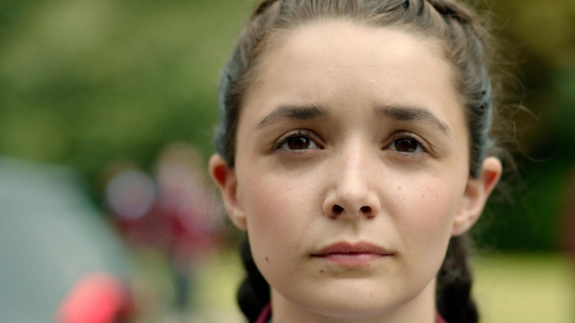 In einer Nahaufnahme sieht man Addie, wie sie mit geflochtenen Zöpfen frontal zur Kamera steht.