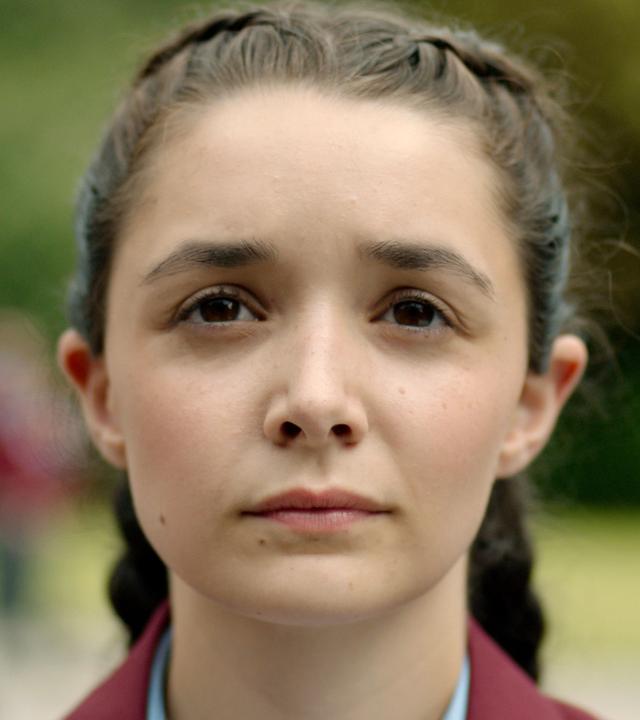 In einer Nahaufnahme sieht man Addie, wie sie mit geflochtenen Zöpfen frontal zur Kamera steht.