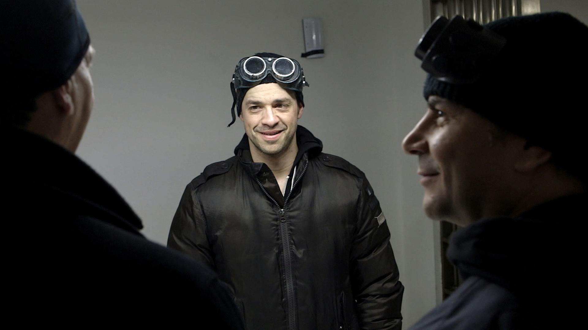 "Hightech-Gangster: Diamanten, Gold und Luxusautos": Drei Männer stehen in einem Raum und sehen sich an. Zwei sind von hinten zu sehen, einer von vorn. Zwei tragen Kappen mit Schutzgläsern, einer nur eine Kappe. Der frontal zu sehende Mann lächelt und schaut den von hinten zu sehenden linken Mann an.