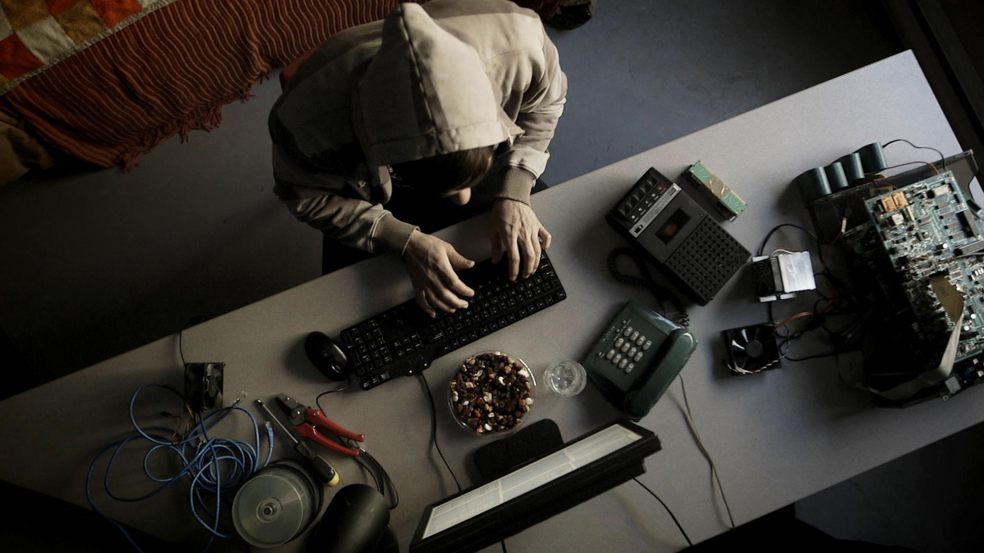 "Hightech Gangster - Ein Computergenie und der Secret Service": Blick von oben: Ein Mann in Kapuzenpullover sitzt an einem Tisch und schreibt etwas auf einer Tastatur. Um ihn herum befinden sich verschiedene Geräte und Gegenstände - wie ein Telefon, ein Monitor, ein Diktiergerät, Kabel, eine Zange und eine CD-Spindel.