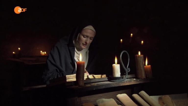 Hildegart von Bingen schreibt etwas an ihrem Schreibtisch, der mit Kerzen beleuchtet ist