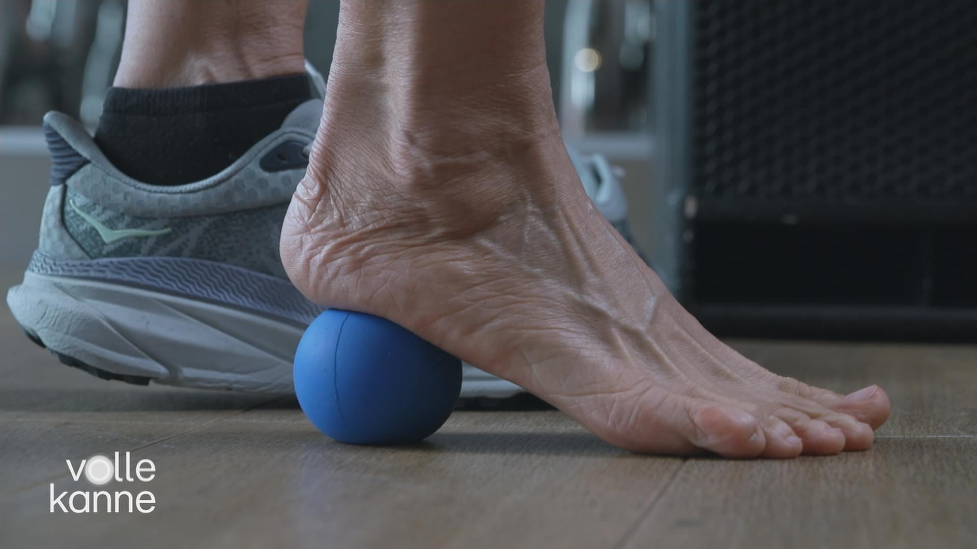 Es sind zwei Füße zu sehen. Der rechte Fuß ist nackt und rollt mit der Ferse auf einem blauen Ball ab. Der linke Fuß ist in einem grauen Schuh.