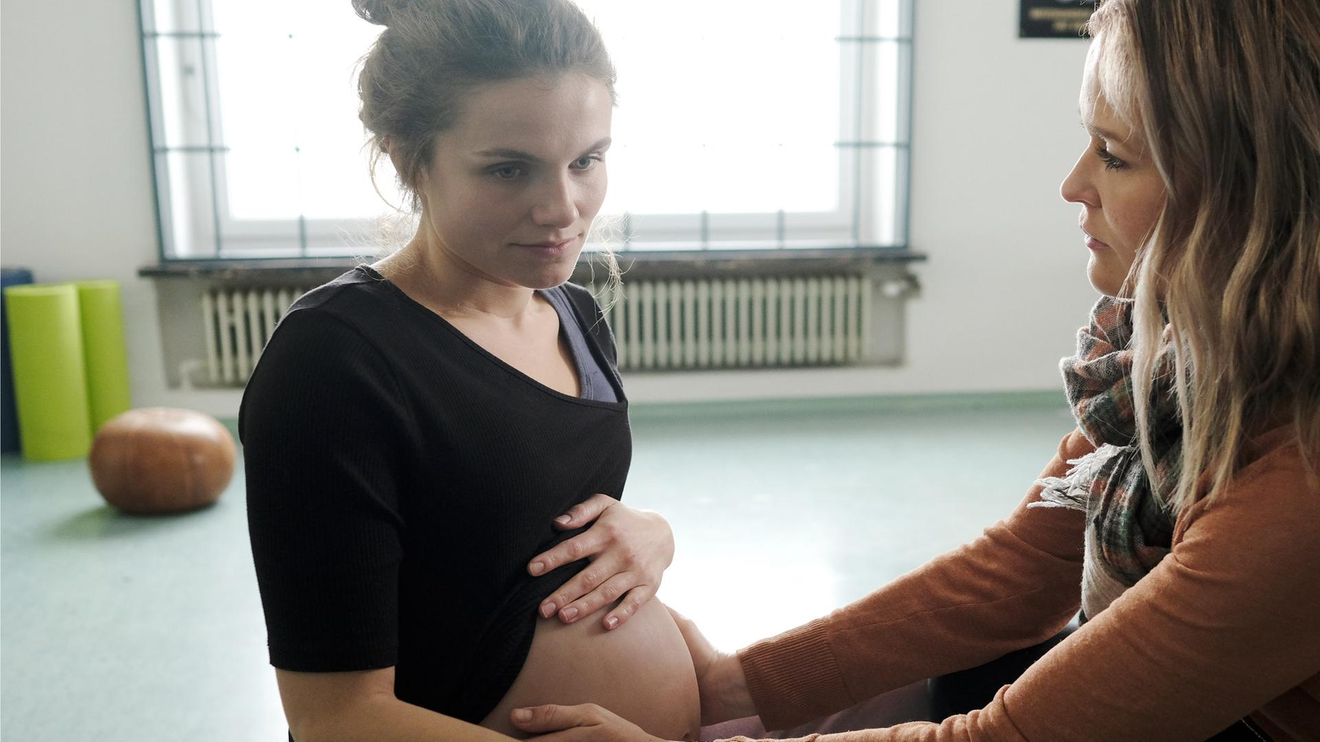  In einem Gemeinschaftsraum sitzen sich Sally Wilkens (Henriette Hölzel) und Lena Lorenz (Judith Hoersch) gegenüber. Lena tastet Sallys Schwangerschaftsbauch ab.