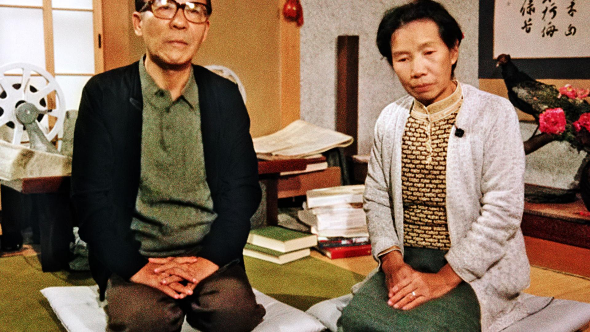 Ein japanischer Mann (links) und eine Frau (rechts) sitzen mit gefalteten Händen auf dem Boden und blicken in die Kamera.