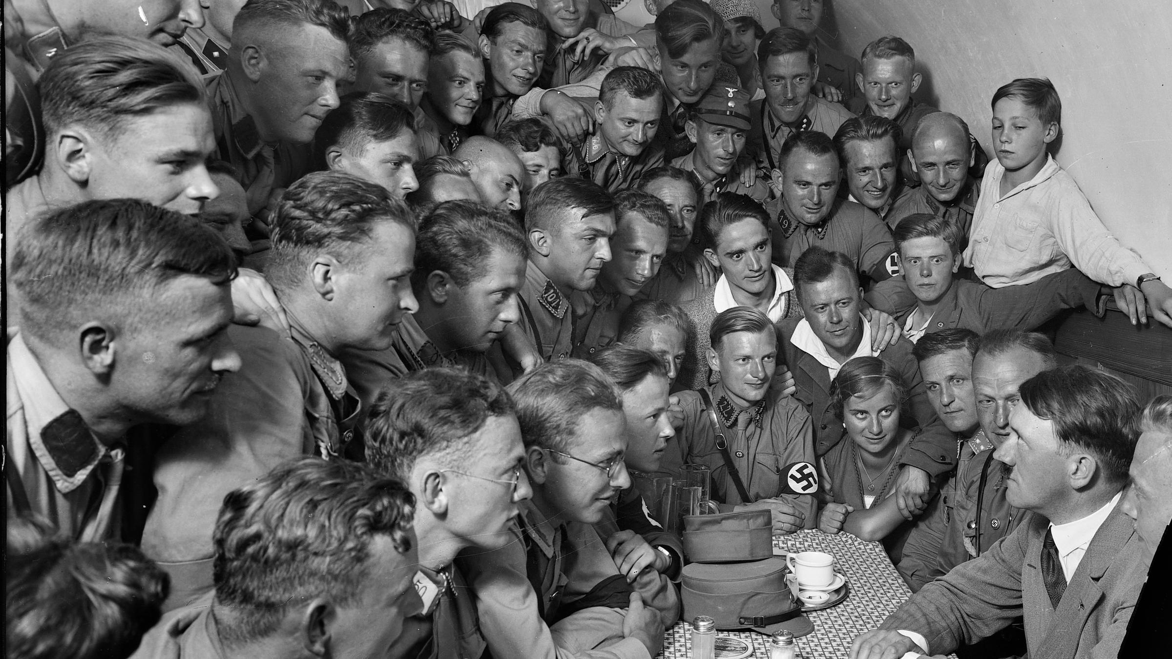 "Hitlers Macht - Der Aufsteiger": Schwarz-Weiß-Foto (Bildausschnitt), das Hitler an einem Tisch sitzend zeigt. Er wird umringt von zahlreichen Menschen.