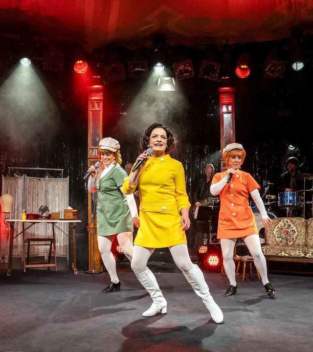 Auf dem Bild sind drei Frauen auf einer Bühne zu sehen, die eine Aufführung darbieten. Die Frauen tragen auffällige, farbenfrohe Kostüme: eine in einem leuchtend gelben Kleid, eine in grün und eine in orange, jeweils mit weißen Strumpfhosen und schwarzen Schuhen. Die Frau in Gelb steht im Vordergrund und hält ein Mikrofon, während die anderen beiden leicht im Hintergrund stehen und ebenfalls Mikrofone halten.   Im Hintergrund ist eine Bühne mit einer Lichtershow zu sehen, die mit roten und weißen Scheinwerfern beleuchtet ist. Der Bühnenboden ist schwarz und es gibt eine verhangene Kulisse aus glitzerndem Material hinter den Darstellerinnen. Eine einfache, retro angelehnte Couch steht seitlich auf der Bühne. An weiteren Bühnendetails erkennt man einige Musikinstrumente, und es gibt einen leicht nebligen Effekt in der Luft, der eine festliche Stimmung erzeugt.