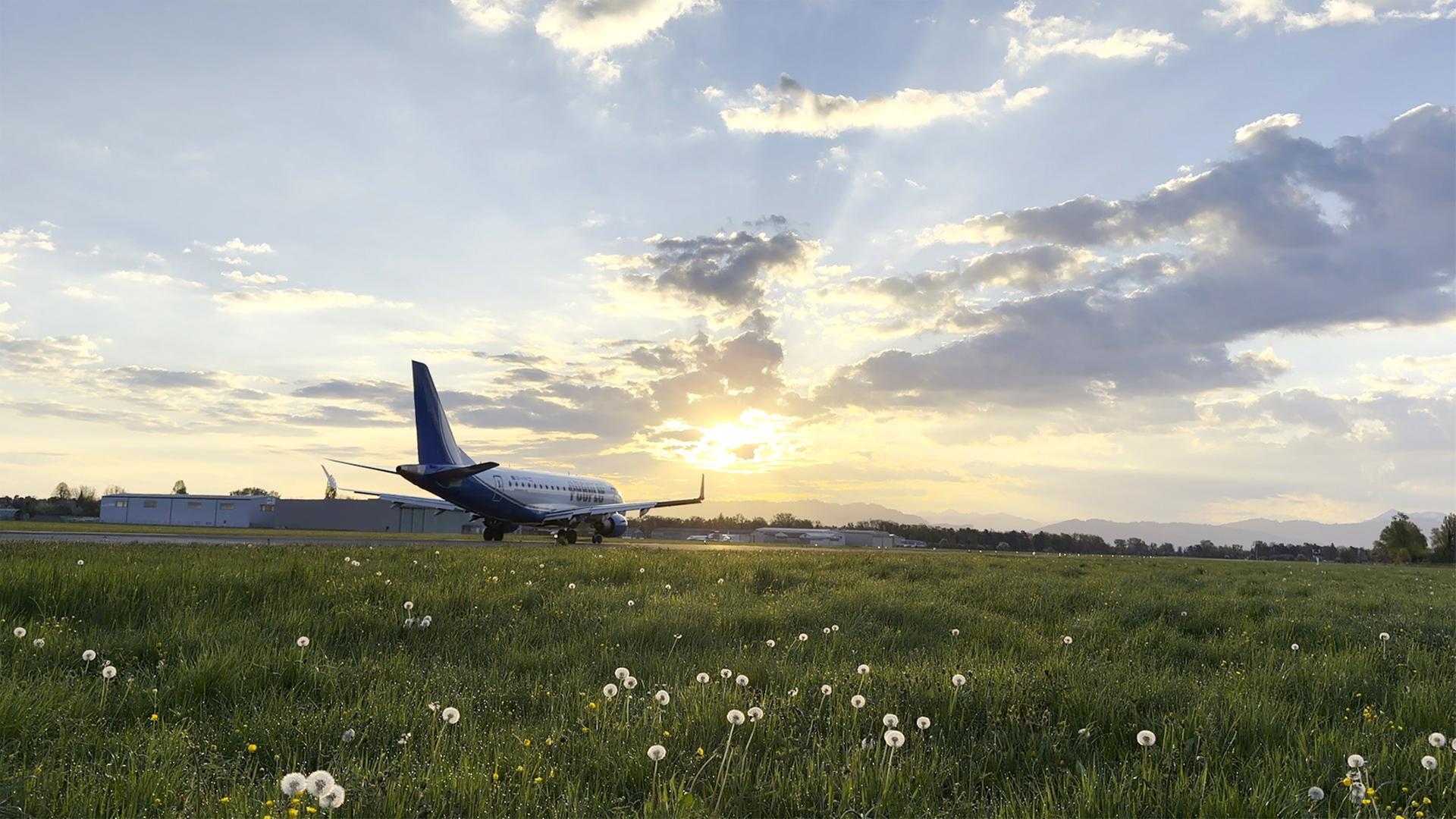 Das Bild zeigt den Flugplatz St. Gallen-Altenrhein in der Schweiz, während der Sonne untergeht. Im Vordergrund ist eine große, grüne Wiese zu sehen, die mit weißen Löwenzahn-Pflanzen übersät ist. Das Gras wirkt frisch und feucht. Rechts im Bild steht ein Flugzeug der Peoples Airline, das sich auf der Landebahn befindet. Der Flugzeugrumpf ist überwiegend blau, und die Tragflächen sind leicht geneigt.   Im Hintergrund sind einige Gebäude und weitere Flugbetriebsinfrastruktur sichtbar, die den Flugplatz ergänzen. Der Himmel ist mit mehreren weißen Wolken durchzogen und schimmert in warmen Gold- und Orangetönen, die durch das Licht der untergehenden Sonne erzeugt werden. Die Landschaft ist sanft und hügelig, mit Bergen, die in der Ferne sichtbar sind.   Die gesamte Szenerie vermittelt eine ruhige und friedliche Atmosphäre, in der die Aktivitäten des Flugplatzes und die natürliche Umgebung harmonisch verbunden sind.
