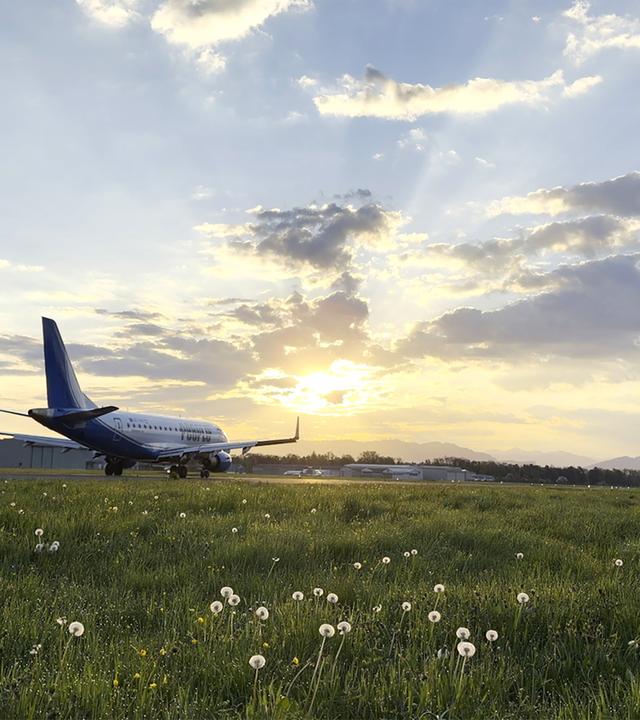 Das Bild zeigt den Flugplatz St. Gallen-Altenrhein in der Schweiz, während der Sonne untergeht. Im Vordergrund ist eine große, grüne Wiese zu sehen, die mit weißen Löwenzahn-Pflanzen übersät ist. Das Gras wirkt frisch und feucht. Rechts im Bild steht ein Flugzeug der Peoples Airline, das sich auf der Landebahn befindet. Der Flugzeugrumpf ist überwiegend blau, und die Tragflächen sind leicht geneigt.   Im Hintergrund sind einige Gebäude und weitere Flugbetriebsinfrastruktur sichtbar, die den Flugplatz ergänzen. Der Himmel ist mit mehreren weißen Wolken durchzogen und schimmert in warmen Gold- und Orangetönen, die durch das Licht der untergehenden Sonne erzeugt werden. Die Landschaft ist sanft und hügelig, mit Bergen, die in der Ferne sichtbar sind.   Die gesamte Szenerie vermittelt eine ruhige und friedliche Atmosphäre, in der die Aktivitäten des Flugplatzes und die natürliche Umgebung harmonisch verbunden sind.